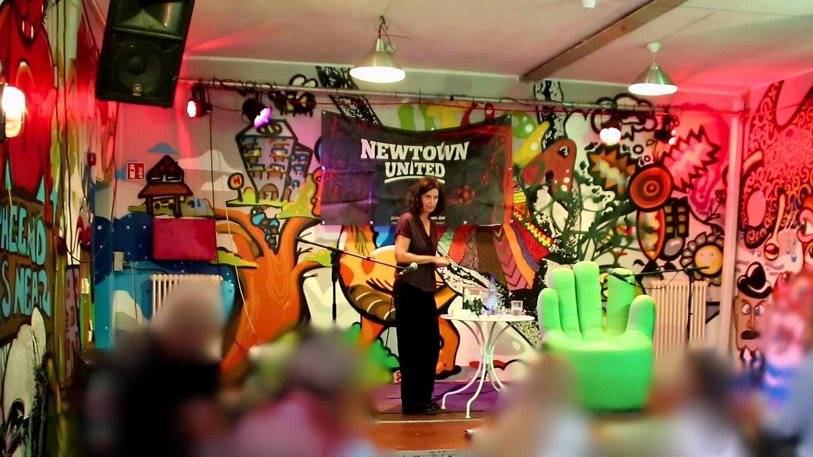 Eva Kranenburg liest vor Publikum im Saal, im Hintergrund das Banner von Newtown United